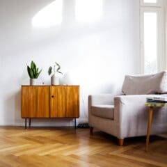 A minimalist living room features a beige armchair with a wooden side table holding a few books. A wooden cabinet with potted plants and a globe lamp sits against a white wall. The room has parquet flooring accented by soft carpets, while natural light brightens the space through large windows.