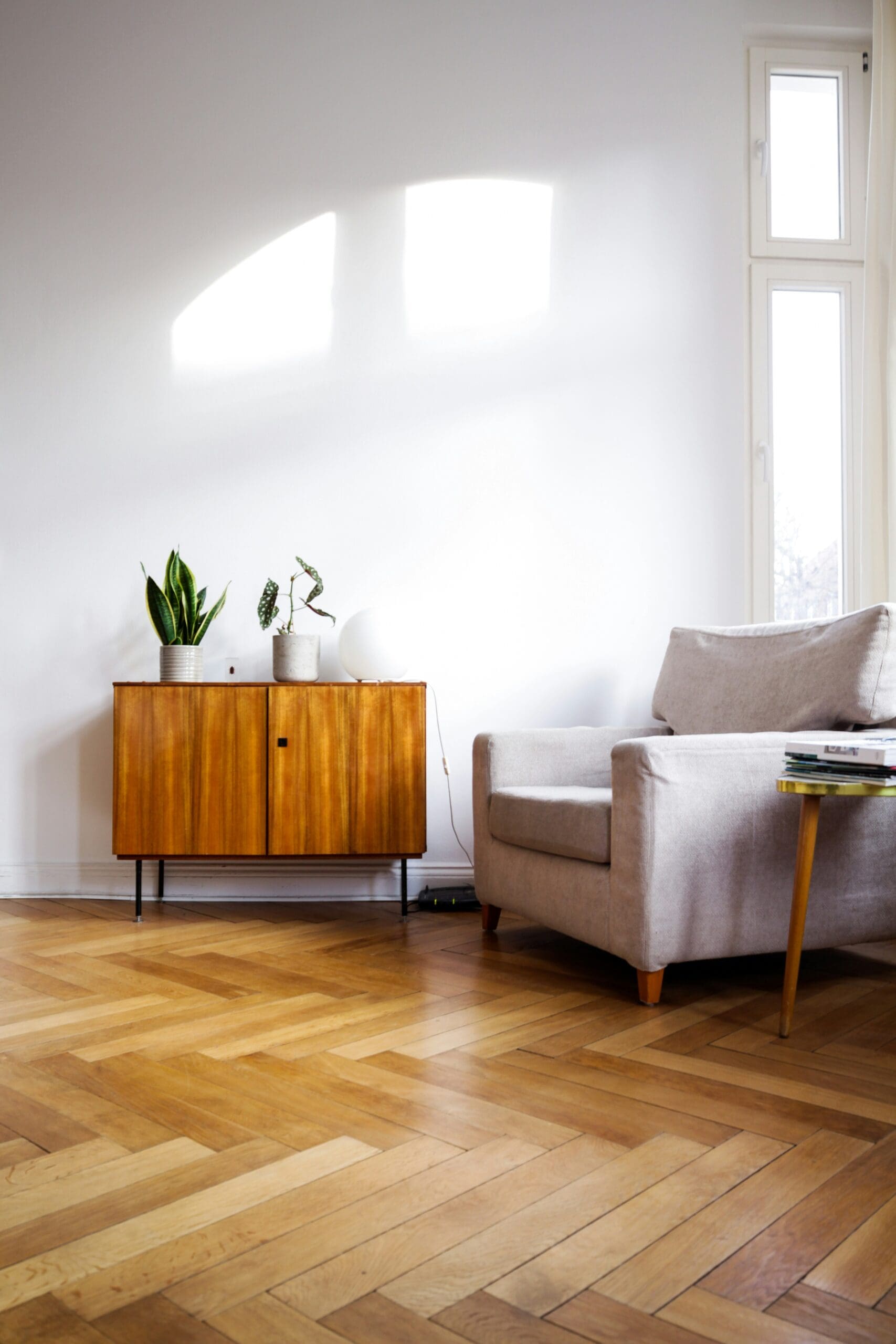 A minimalist living room features a beige armchair with a wooden side table holding a few books. A wooden cabinet with potted plants and a globe lamp sits against a white wall. The room has parquet flooring accented by soft carpets, while natural light brightens the space through large windows.