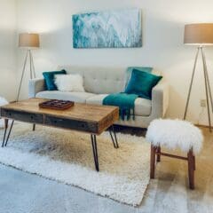 A cozy living room features a light-colored sofa with teal pillows and a throw blanket. A wooden coffee table with a game board sits on white shag carpets. Two fluffy stools and modern lamps with beige shades flank the setup. An abstract painting hangs on the wall.