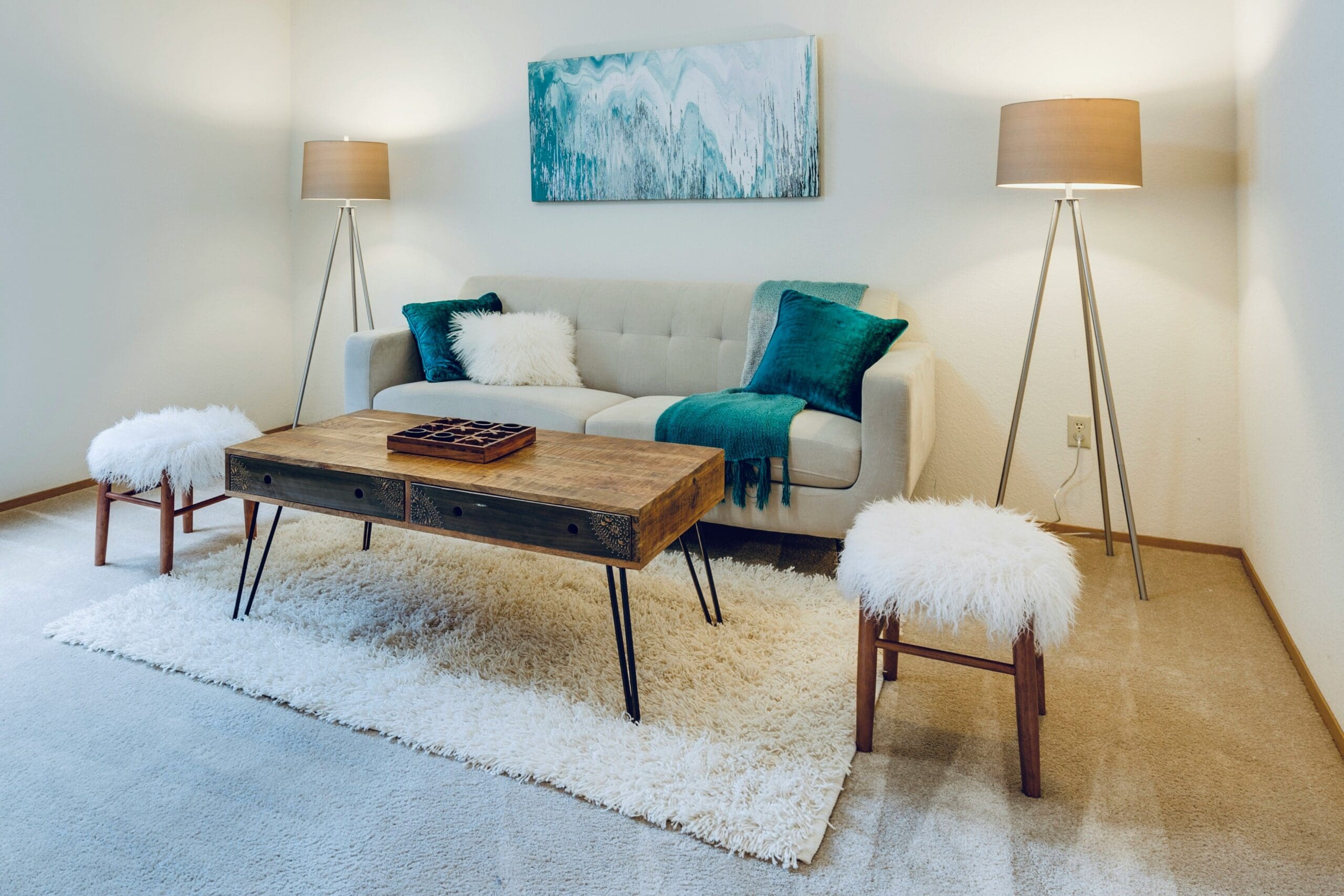 A cozy living room features a light-colored sofa with teal pillows and a throw blanket. A wooden coffee table with a game board sits on white shag carpets. Two fluffy stools and modern lamps with beige shades flank the setup. An abstract painting hangs on the wall.