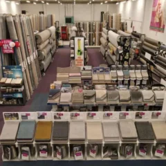 A wide-angle view of a carpet store showcases rows of various carpet rolls standing vertically. Displays of carpet samples are arranged on racks in the center. The store is well-lit with aisles for easy browsing. Sale signs and customer service desks are visible, highlighting the vast selection of carpets.