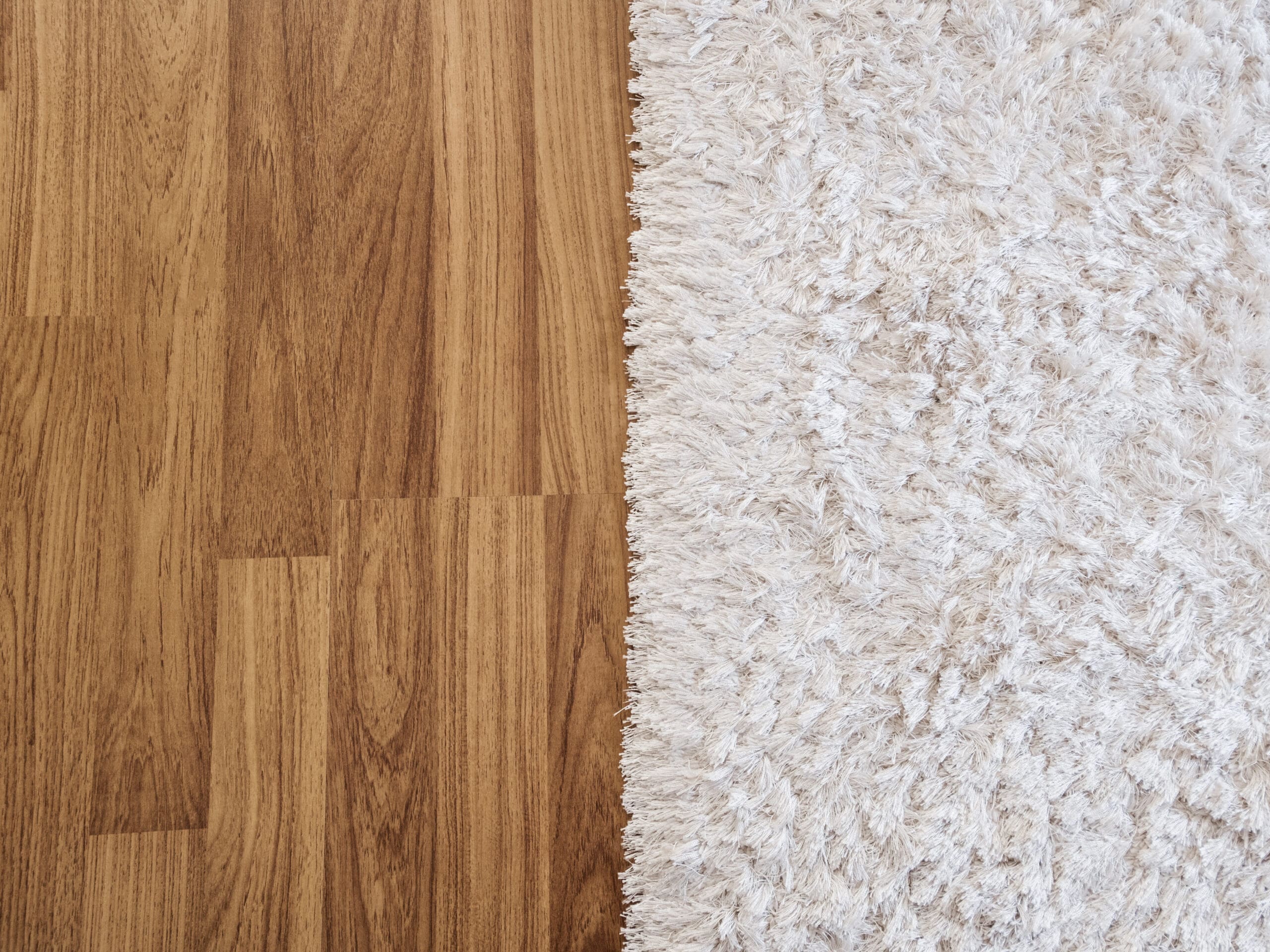 Close-up image of a soft, white, fluffy rug partially covering a wooden floor with a medium brown finish. The rug is on the right side, and the wooden floor is on the left, creating a contrast in textures and colors.