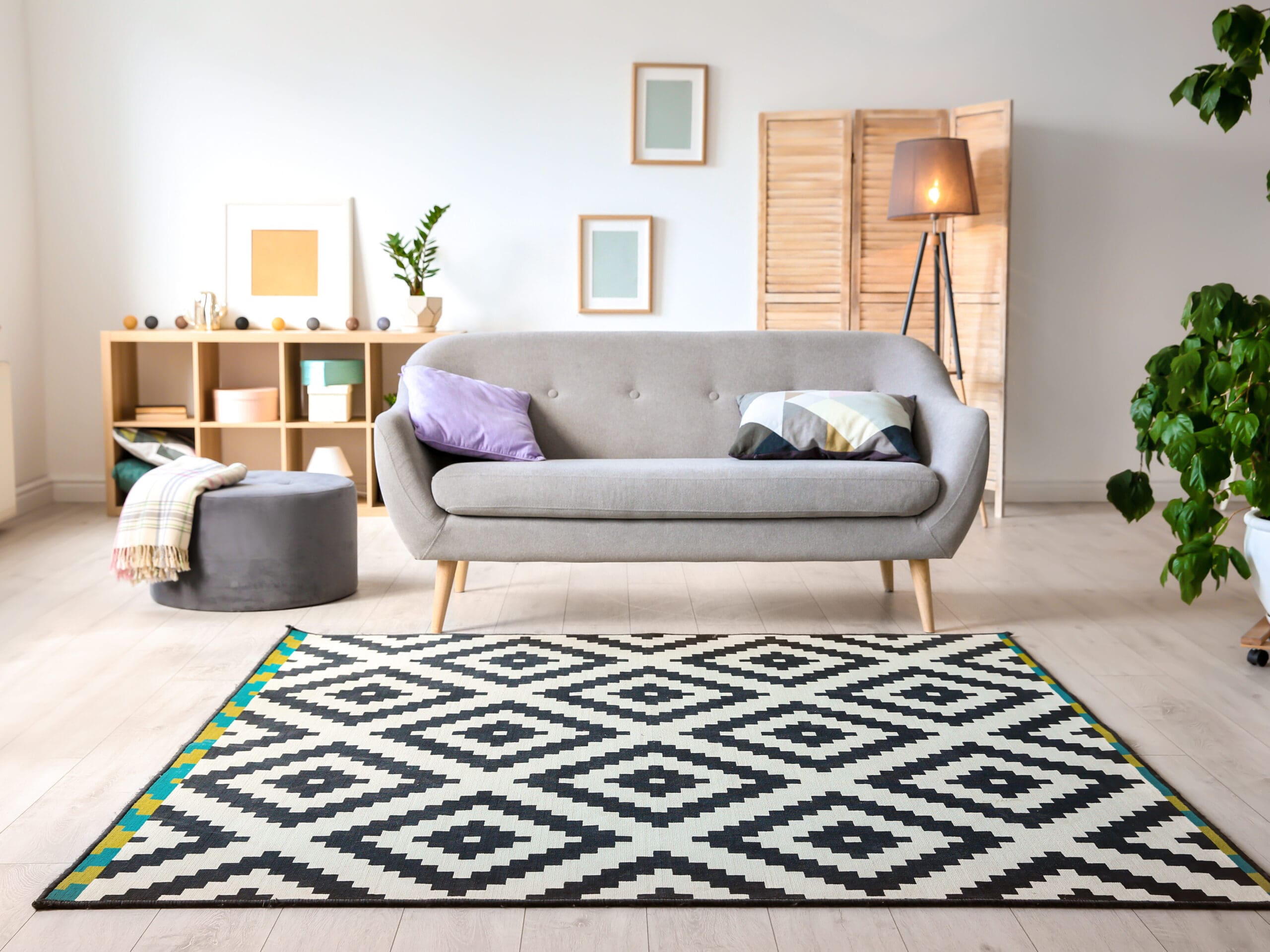 A modern living room interior featuring a gray sofa with purple and white pillows, a gray ottoman, a geometric patterned black and white rug, artwork on the walls, a wooden screen, a floor lamp, and a large green plant in the corner.