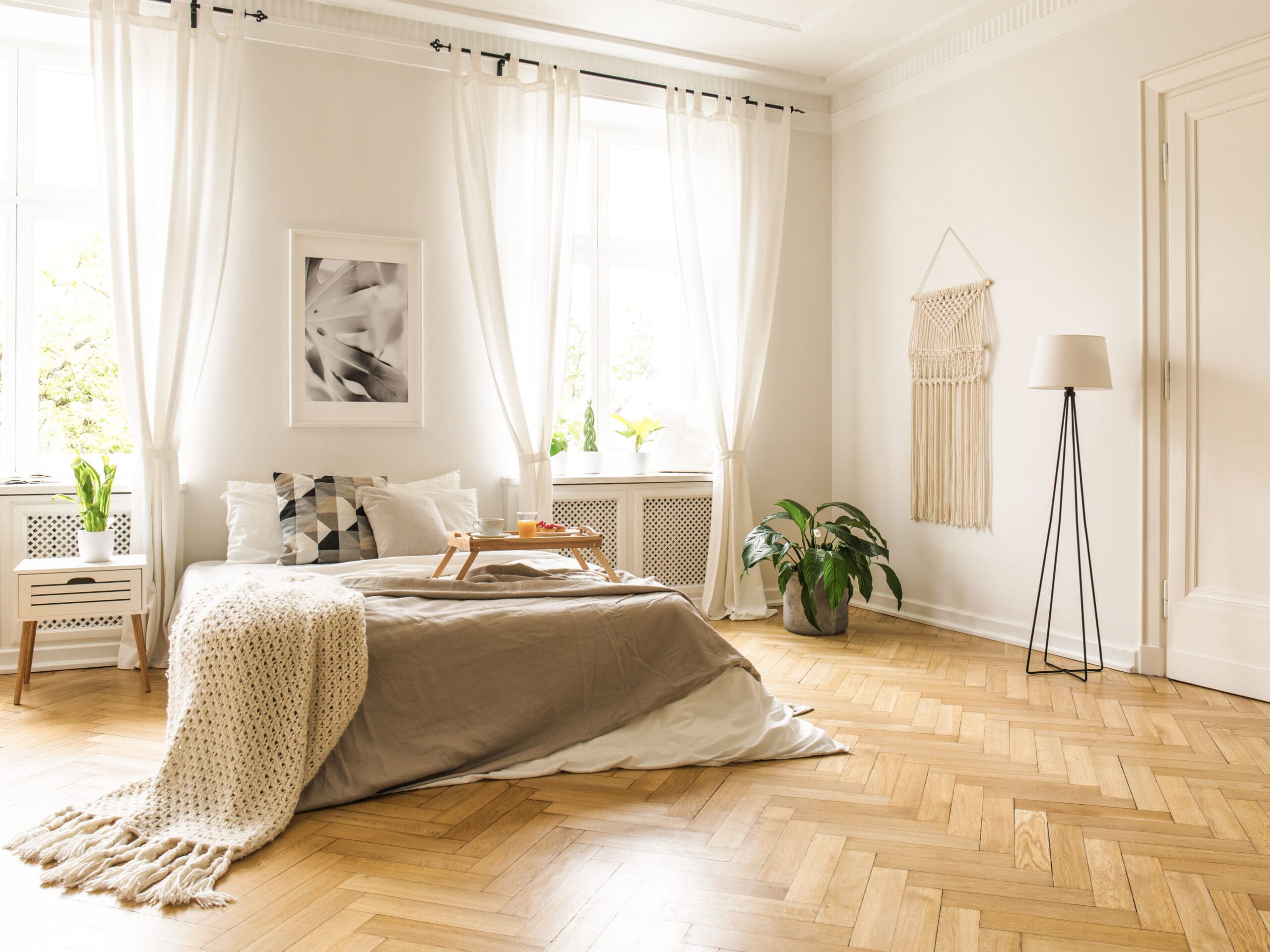 A bright, minimalist bedroom with a neatly made bed featuring neutral-colored bedding and a knitted blanket. Sunlight streams through large windows with sheer curtains. A plant, floor lamp, and wall art add simple decor while a breakfast tray sits on the bed.