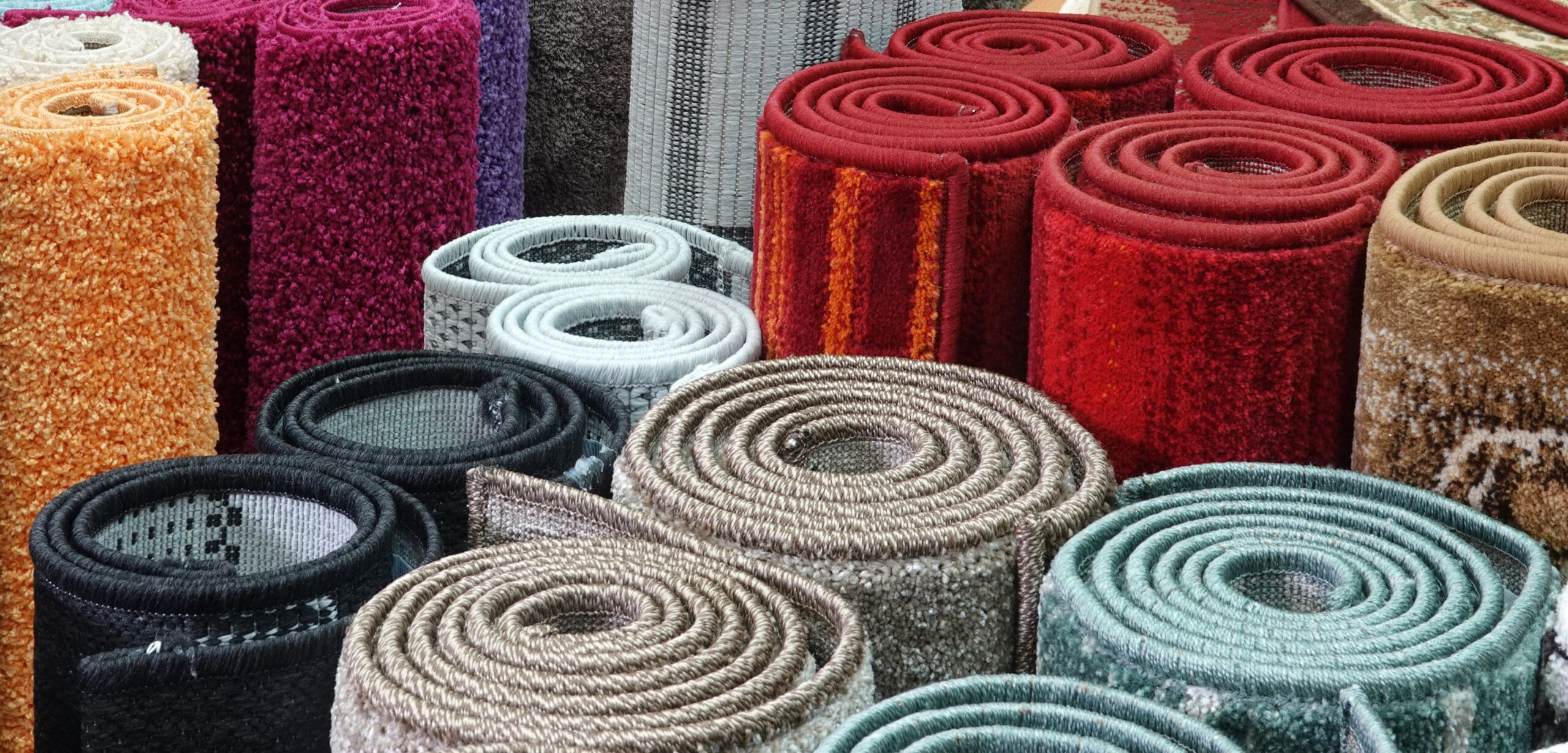 A variety of rolled-up carpets are displayed upright in different colors and textures, including shades of red, orange, green, brown, black, and white. Some carpets are plain while others have patterns or textured surfaces.