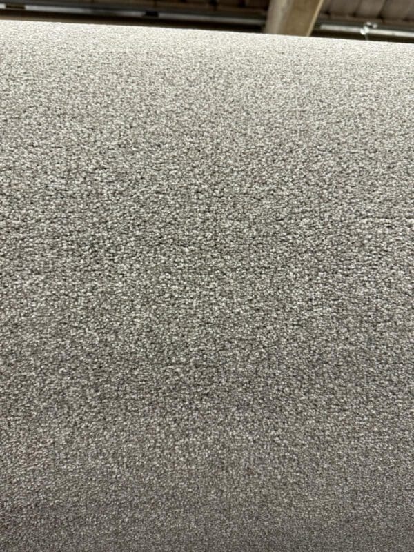 A close-up view of a large roll of grey carpet, showing the texture of the dense, small loops of fiber. The background includes some blurred warehouse or factory features.