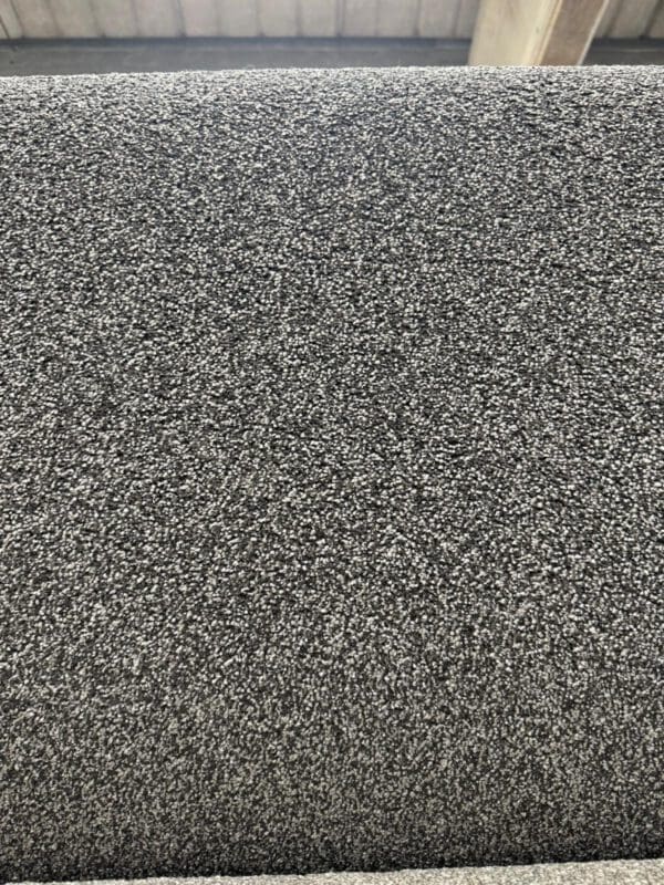 A close-up image of a roll of grey carpet with a speckled texture, showcasing the material's pattern and density. The background includes part of the ceiling with wooden beams, suggesting the photo was taken in a warehouse or store.