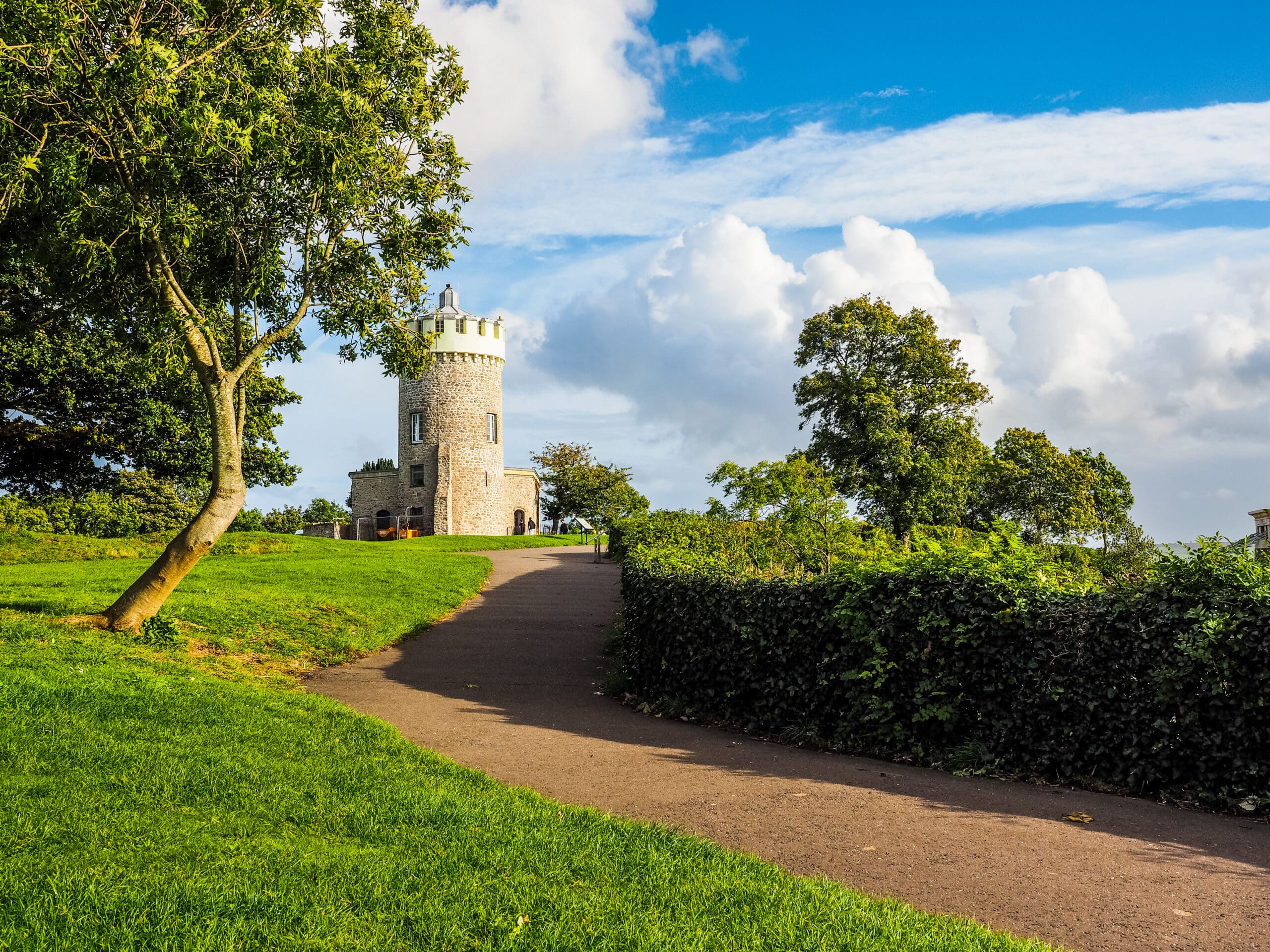 A scenic view of a stone tower with a turret surrounded by lush green trees and grass. A winding pathway leads up to the tower, and the sky is clear with a few scattered clouds. The setting appears to be a peaceful park or garden.