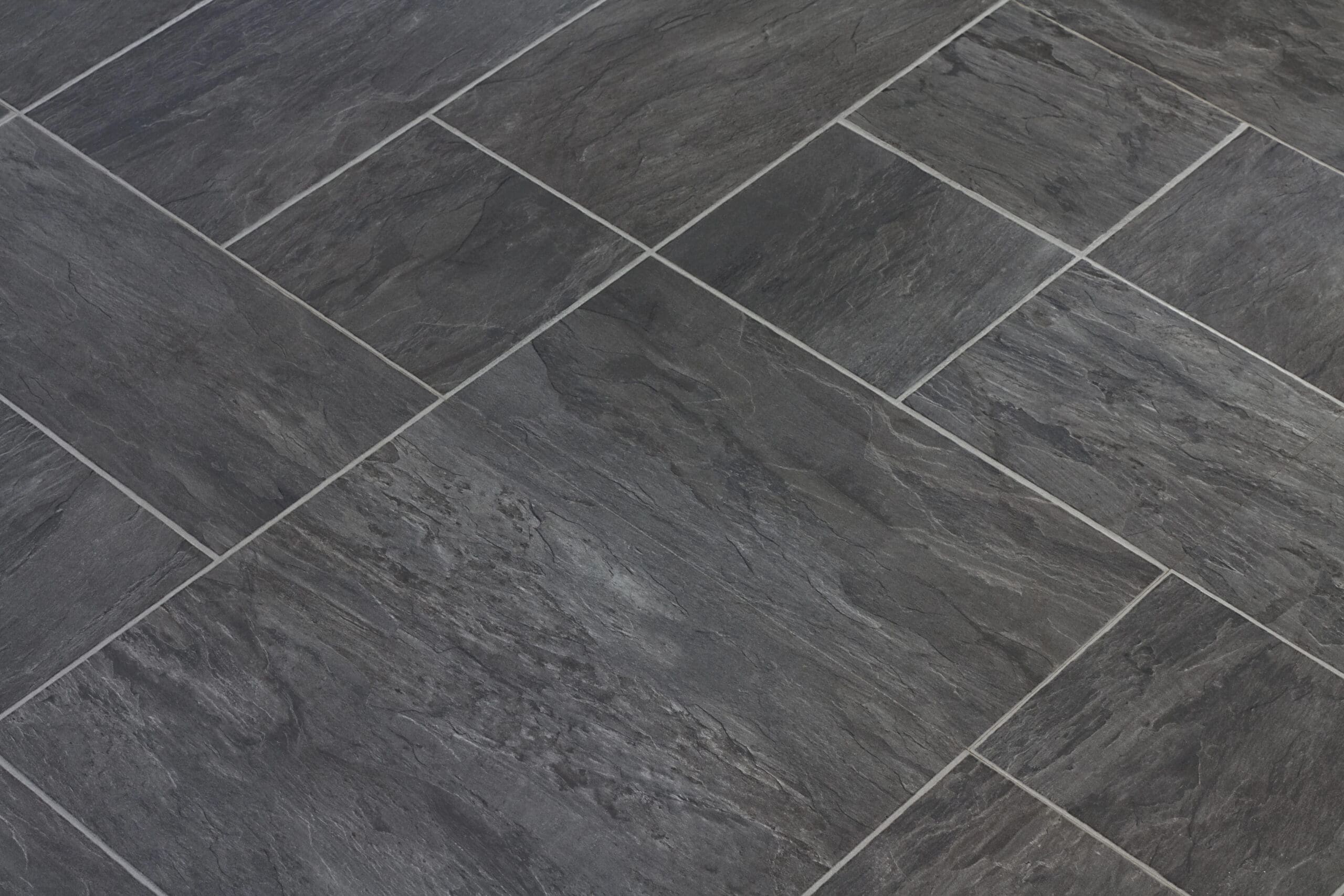 A close-up image of a floor made of dark gray slate tiles arranged in a grid pattern. The tiles have a natural, textured stone appearance with subtle variations in shade and texture, giving the floor a sleek and modern look.