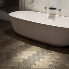 A modern bathroom features a free-standing white oval bathtub placed against a white marble wall with sleek fixtures. The floor is covered with dark, herringbone-patterned tiles, creating a stylish and contemporary feel.