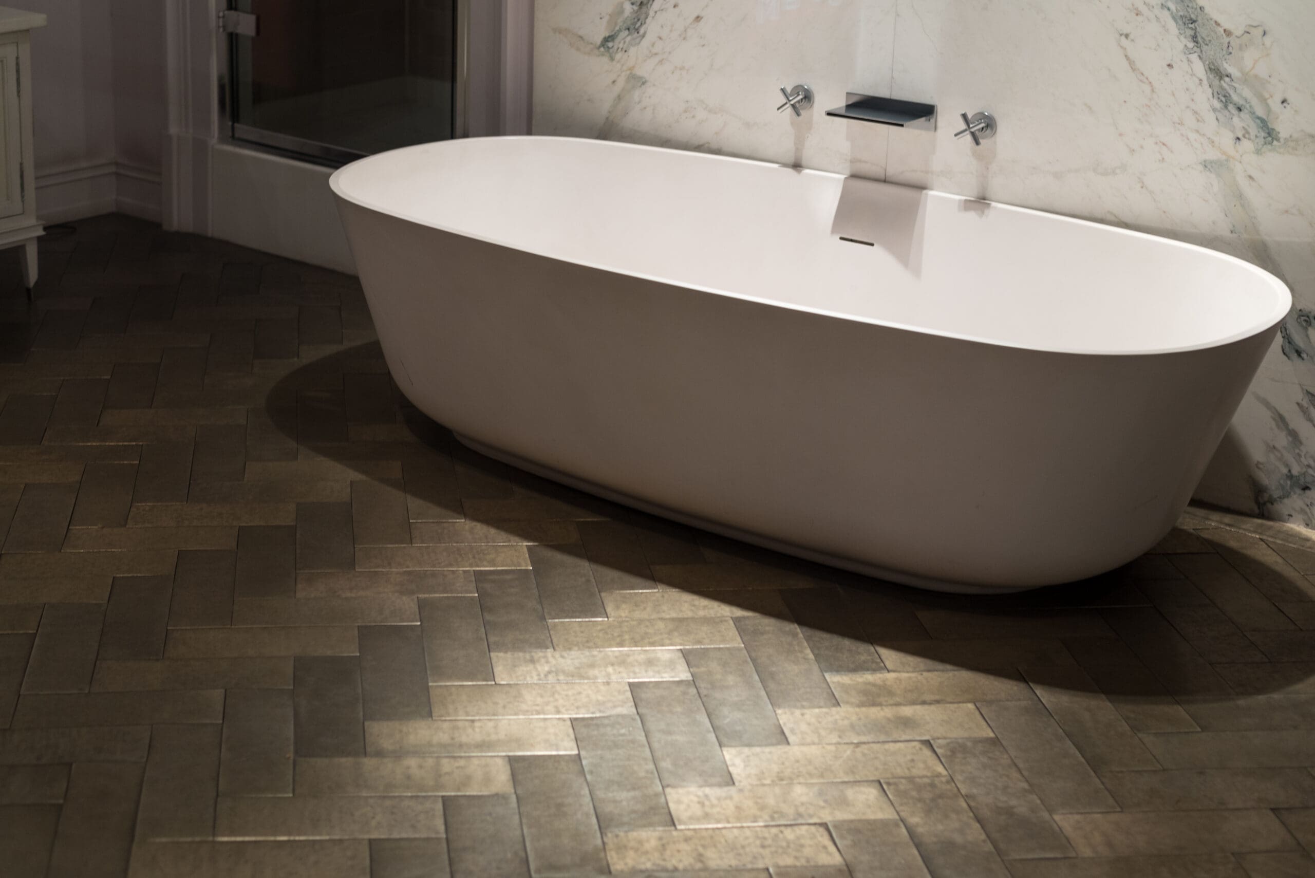 A modern bathroom features a free-standing white oval bathtub placed against a white marble wall with sleek fixtures. The floor is covered with dark, herringbone-patterned tiles, creating a stylish and contemporary feel.