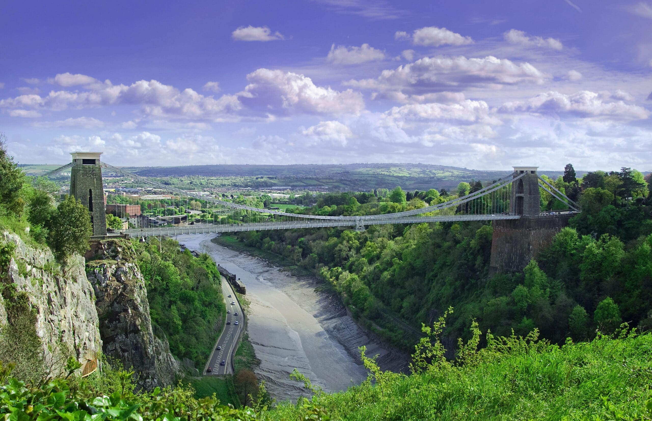 A wide view of the Clifton Suspension Bridge spanning a deep gorge with a river below, surrounded by lush greenery that carpets the landscape. The sky is bright blue with scattered clouds, and a distant panorama of fields and buildings is visible.