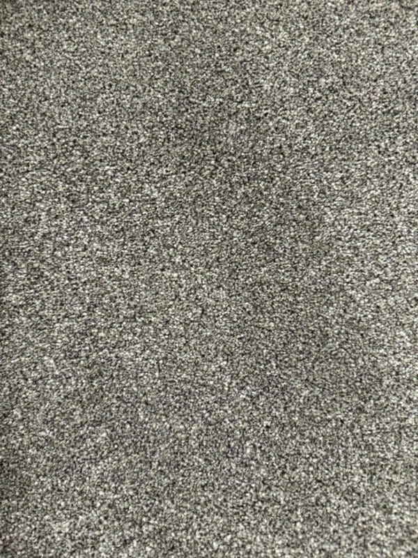 A close-up image of a grey textured carpet floor. The fibers of the carpet are densely packed, creating a uniform and slightly speckled surface. The overall appearance is soft and plush.