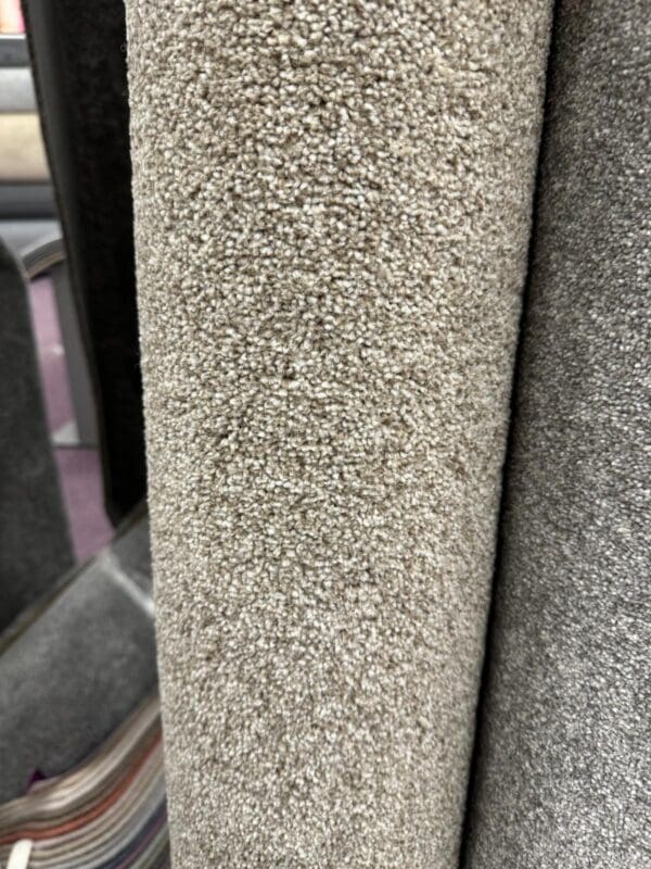 Close-up of several rolled-up carpet samples in various neutral colors, including beige, gray, and a striped pattern. The focus is on the texture and fibers of the central beige carpet, which is the S/F Rustique Country Life 1.7x4m. The background showcases other carpet rolls, partially visible.