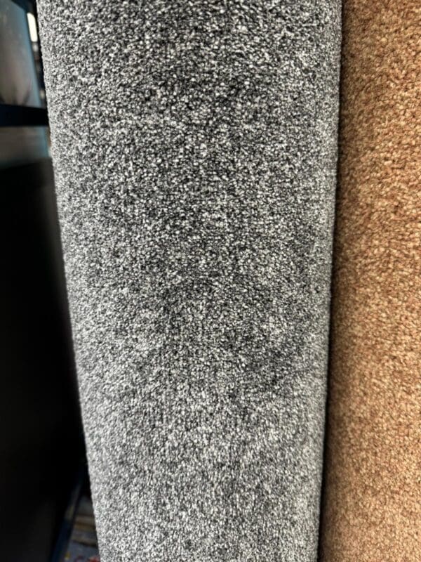 A close-up of two Trident Twilight 2.4x4m carpet rolls standing vertically next to each other. The carpet on the left is gray with a textured surface, and the one on the right is brown. Both carpets appear plush and thick.