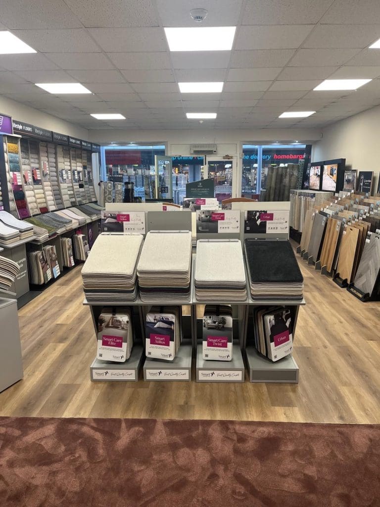 A carpet store interior with various carpet samples displayed on stands. Multiple racks showcase carpet swatches in different colors and textures. Floor is wooden with some carpeted sections. Storefront windows show neighboring shops outside.