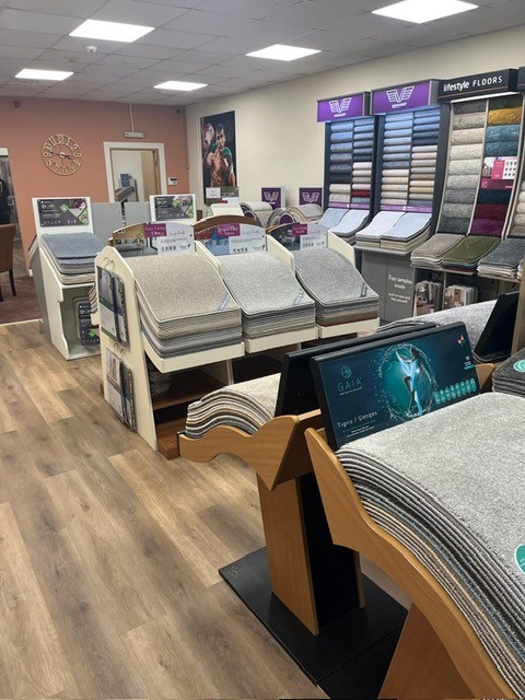 Interior of a carpet and flooring store with multiple displays of carpet samples in various shades of gray and beige. There are rows of samples on wooden stands, racks showcasing flooring options, and promotional signage on the walls.