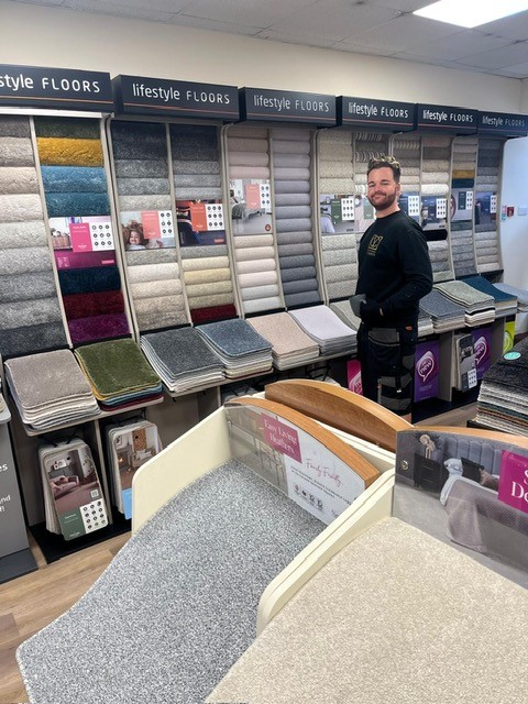 A person stands in a flooring store, surrounded by various carpet samples displayed in racks. They are wearing a black outfit and smiling at the camera. The samples range in color from neutral tones to vibrant shades.