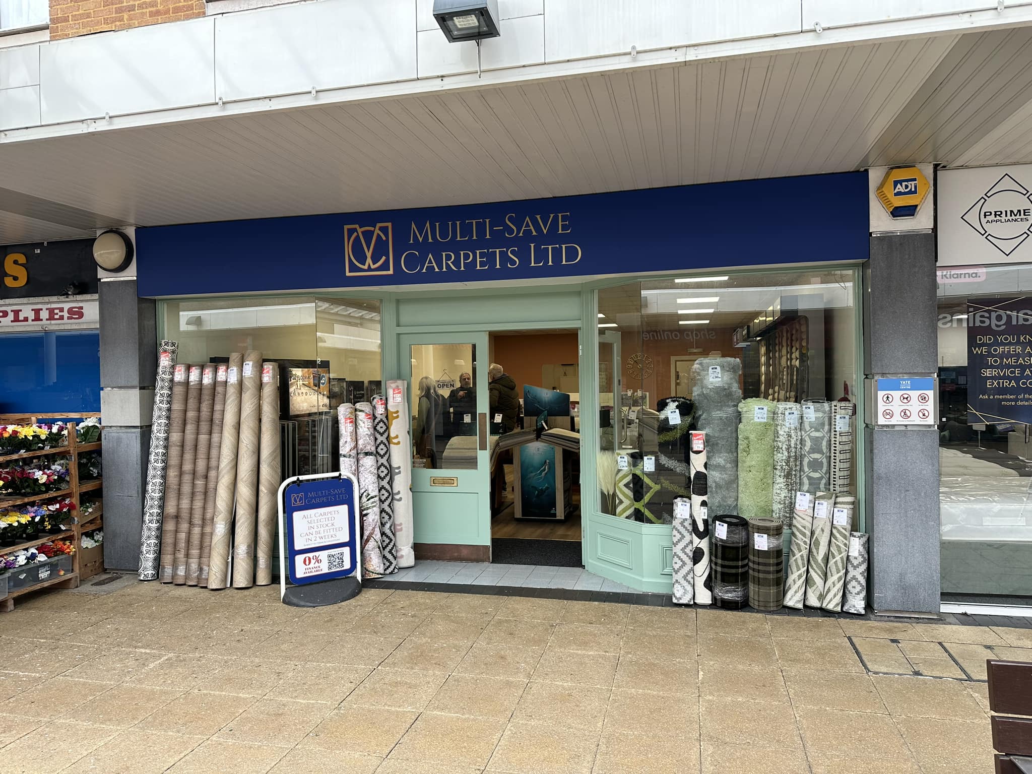 Storefront of "Multi-Save Carpets Ltd" displaying rolled carpets outside. The entrance has a light blue door and window frame. A signboard with offers is near the door, and a neighboring store's produce is partially visible on the left.